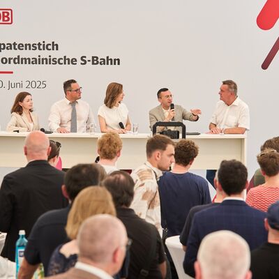 Fünf Menschen stehen auf einer Tribüne. Einer hat ein Mikrofon in der Hand.