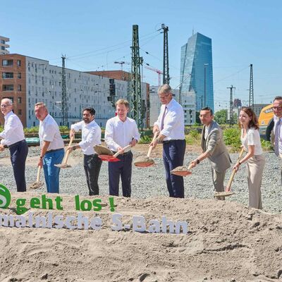 Mehrere Personen stehen auf einem Baufeld. Mit einem Spaten schippen sie Sand auf einen Haufen.