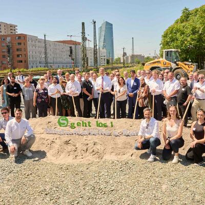 Mehrere Menschen stehen vor einem Sandhaufen. Einige haben einen Spaten in der Hand.