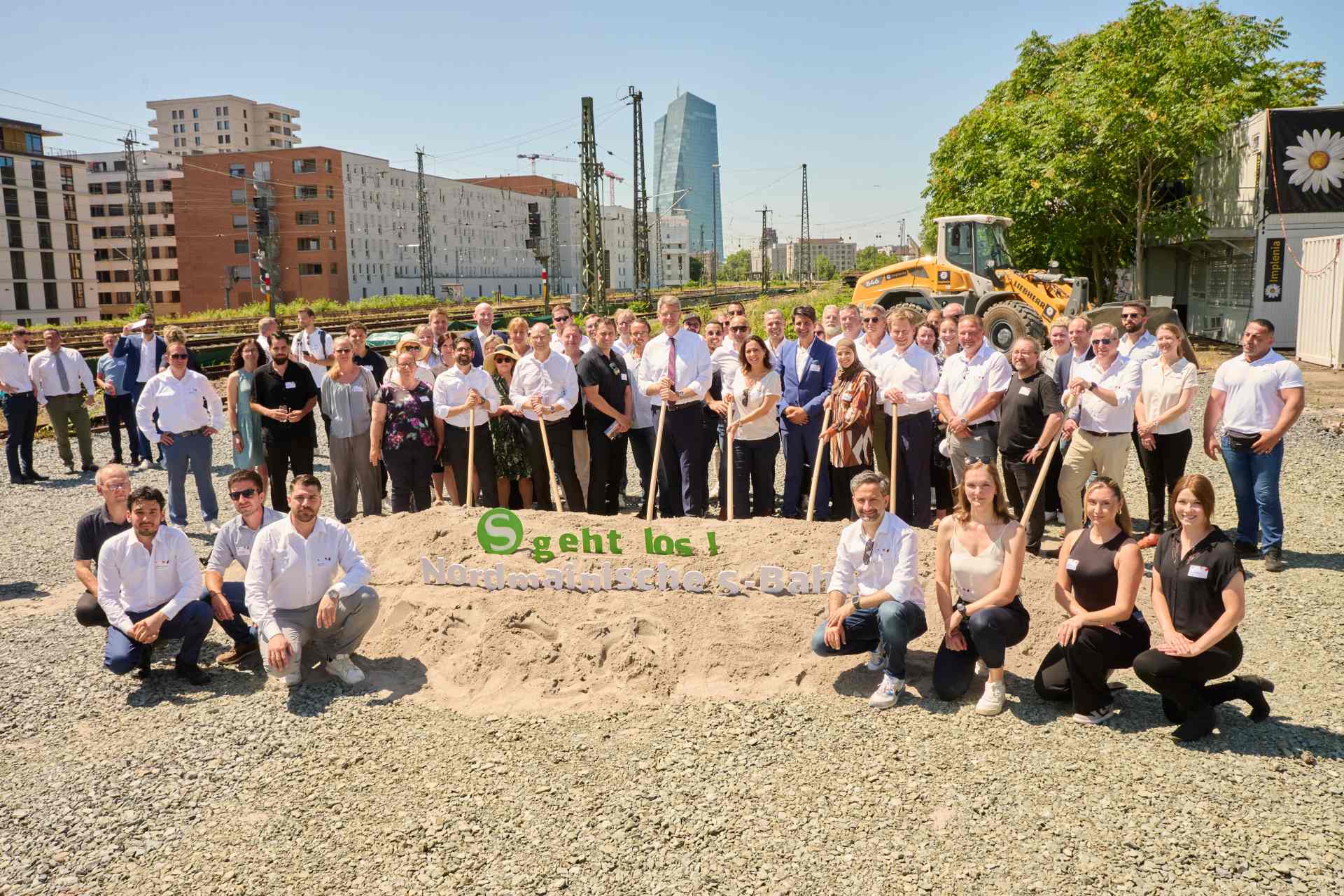 Mehrere Menschen stehen vor einem Sandhaufen. Einige haben einen Spaten in der Hand.