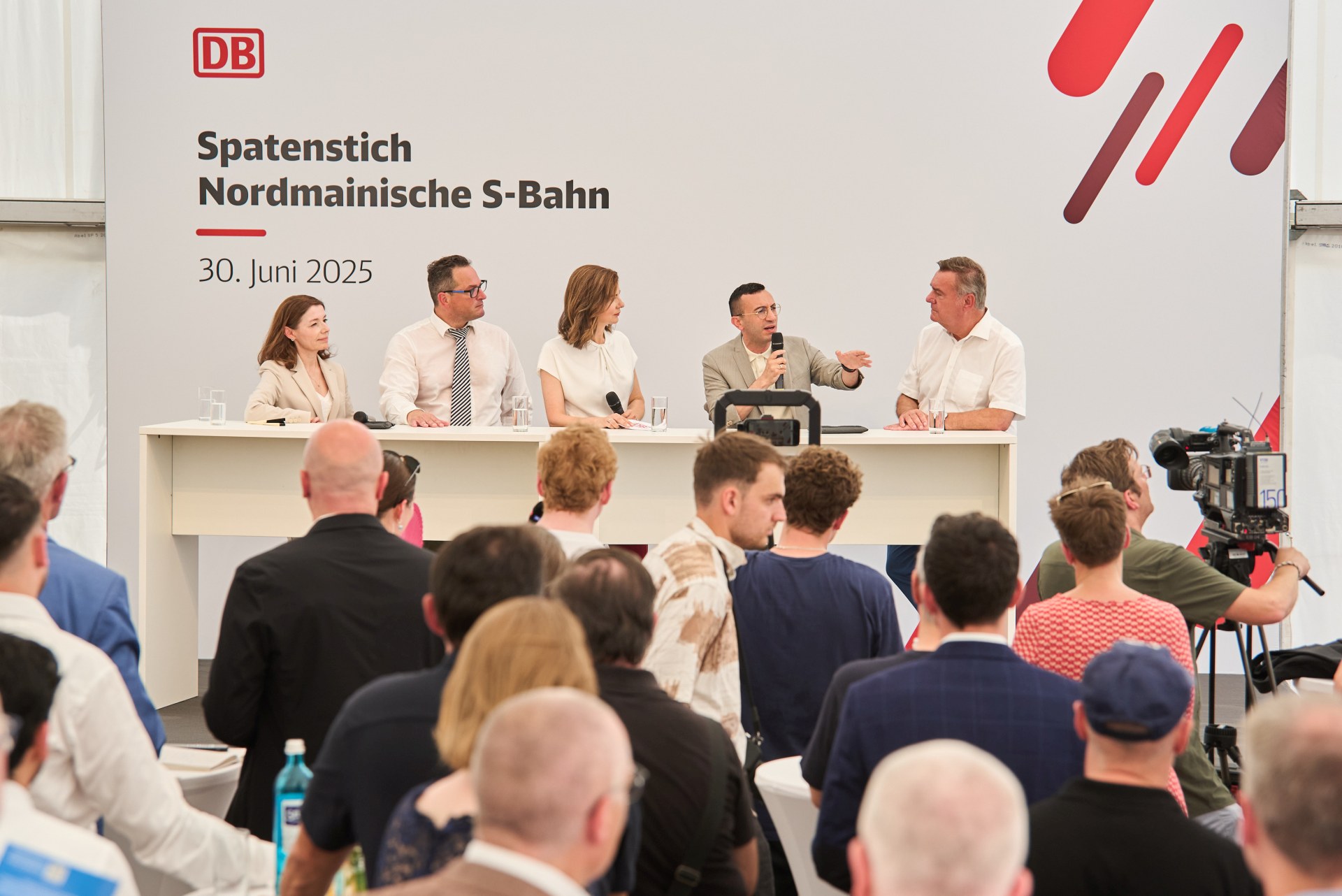 Fünf Menschen stehen auf einer Tribüne. Einer hat ein Mikrofon in der Hand.