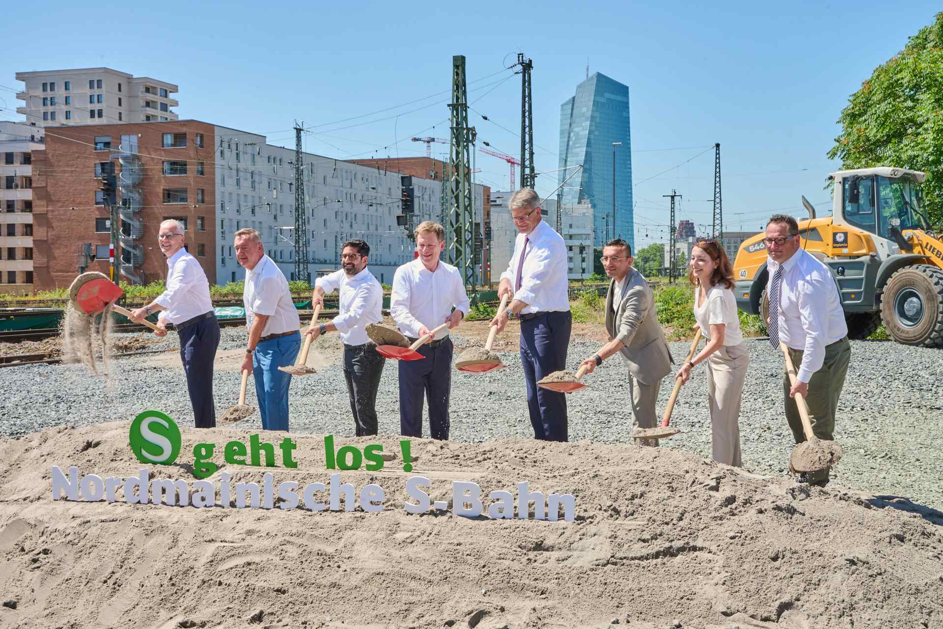 Mehrere Personen stehen auf einem Baufeld. Mit einem Spaten schippen sie Sand auf einen Haufen.