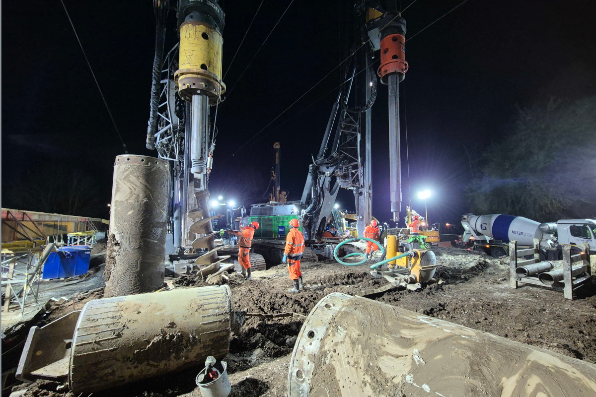 Mitarbeiter inspizieren zwei Bagger mit Bohrpfahlgeräten. Die nötigen weiteren Maschinen stehen um die Baustelle herum.